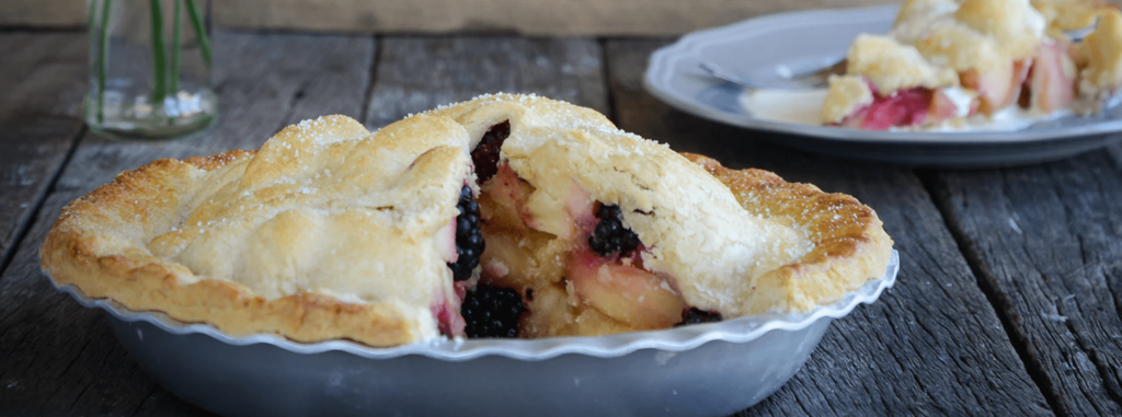 A fruit filled pie sits with a slice cut out in a Silverwood Bakeware Pie tin on a rustic wooden table with a plate beyond with a pie slice on top.
